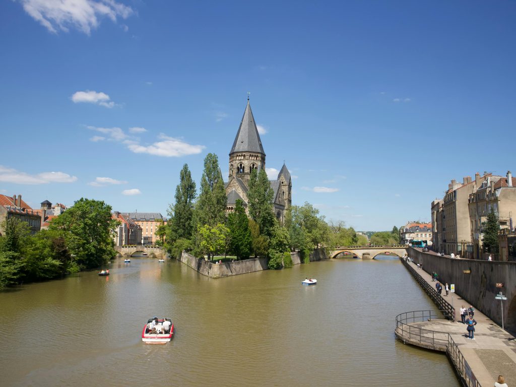 Photo des berges de la Moselle avec d'un cîté les habitations, au milieu une basilique et à gauche la nature